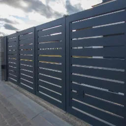 Nowoczesna, przesuwna brama w kolorze antracytu z poziomymi szczelinami, zamontowana na posesji z szarym ogrodzeniem modułowym i kostką brukową.