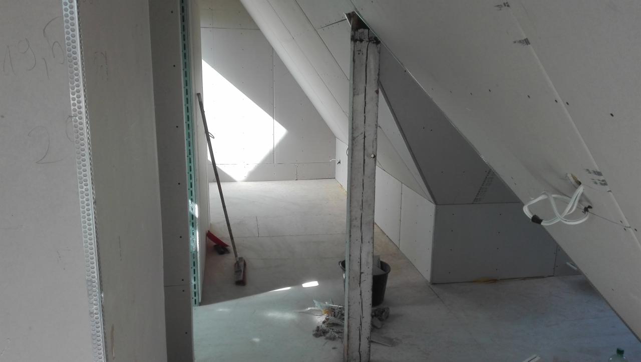 Adaptacja poddasza: widok na pomieszczenie z zamontowanymi płytami na skosach i ścianach, widoczna drewniana belka konstrukcyjna, kabel elektryczny oraz narzędzia i wiadro na podłodze.