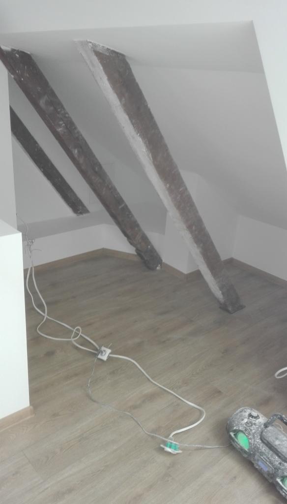 Poddasze z drewnianymi belkami stropowymi i nową podłogą laminowaną, widoczne kable elektryczne i narzędzia, ściany pomalowane na biało.