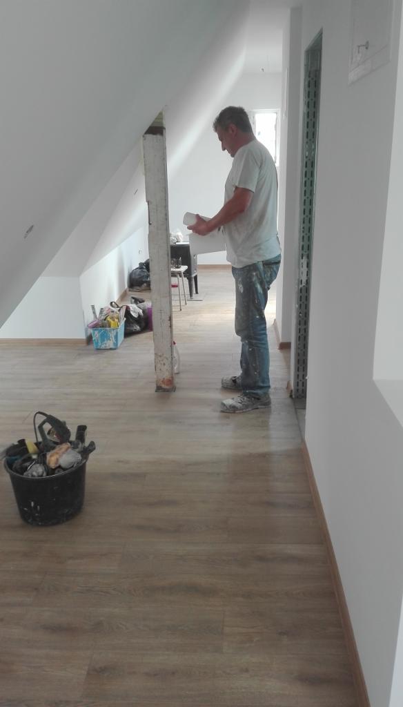 Pracownik w trakcie montażu płyty karton gips na poddaszu, widoczna konstrukcja skośna dachu, przygotowane narzędzia w wiadrze i niezagospodarowana przestrzeń w tle.