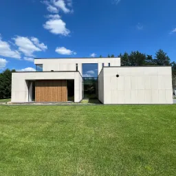 Nowoczesny dom z jasną elewacją i drewnianymi elementami wykończeniowymi, otoczony zielonym trawnikiem i drzewami, pod błękitnym niebem z chmurami.