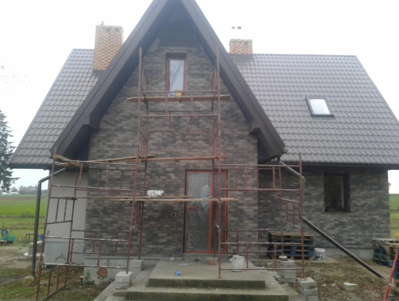 Dom jednorodzinny w trakcie prac wykończeniowych elewacji, widoczne rusztowania, palety z materiałami budowlanymi i fragmenty ocieplenia