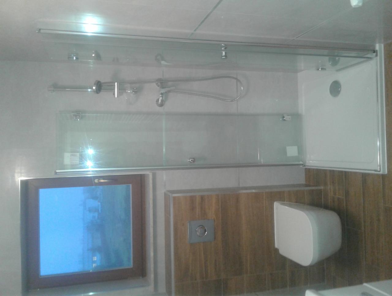 Nowoczesna kabina prysznicowa z przesuwnymi drzwiami, zamontowana w łazience z szarymi płytkami i drewnopodobną zabudową, widoczny fragment okna z niebieskim odbiciem.