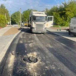 Świeżo wylany asfalt na ulicy z widocznymi studzienkami kanalizacyjnymi, w tle ciężarówka Scania z otwartymi drzwiami, koparka i zaparkowany bus firmy Hemper Bau.