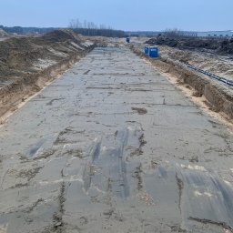 CUDEX - Przygotowany teren pod budowę drogi, z widocznymi śladami maszyn i wyrównaną powierzchnią, w tle zabudowania i linia drzew.