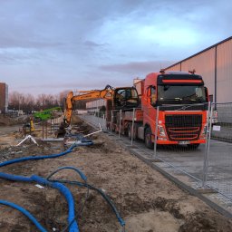 CUDEX - Pomarańczowy ciągnik siodłowy Volvo przewozi żółtą koparkę Hyundai na niskopodwoziowej naczepie, widoczne niebieskie rury na ziemi i teren budowy w tle.