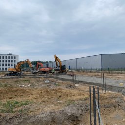 CUDEX - Przygotowanie terenu pod budowę hali: widoczne trzy koparki, w tym jedna marki Hyundai, pracujące na placu budowy z widocznymi fundamentami i elementami zbrojenia, w tle budynek biurowy i hala...