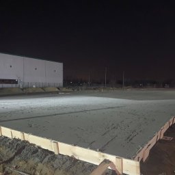 CUDEX - Wylana płyta betonowa w drewnianym szalunku na placu budowy nocą, w tle budynek magazynowy.