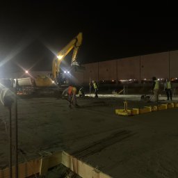 CUDEX - Nocna niwelacja terenu pod budowę, widoczna koparka JCB, pracownicy w kamizelkach odblaskowych i przygotowane zbrojenie.