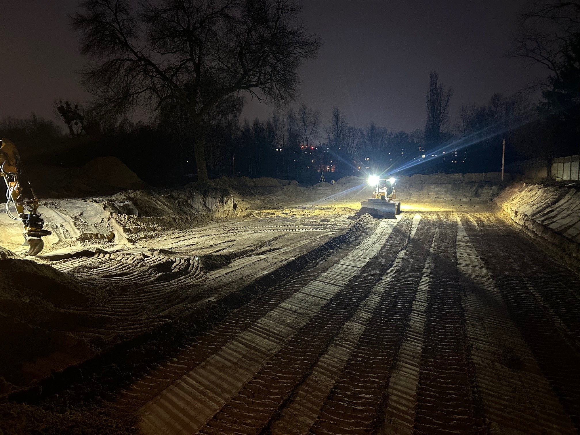 Dwa traktory pracują na placu budowy w nocy, oświetlając teren reflektorami. Wyraźne ślady opon na ziemi, w tle drzewa i budynki.