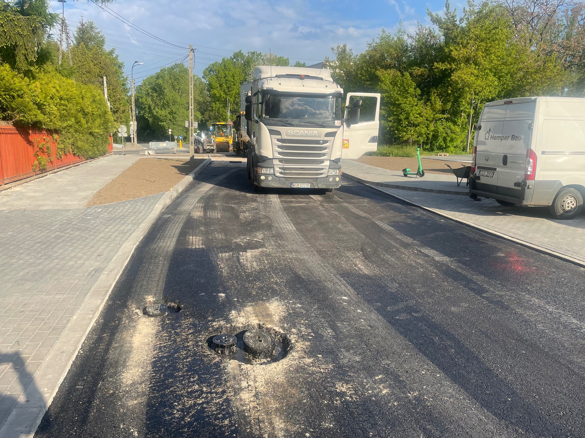 Świeżo wylany asfalt na ulicy z widocznymi studzienkami kanalizacyjnymi, w tle ciężarówka Scania z otwartymi drzwiami, koparka i zaparkowany bus firmy Hemper Bau.