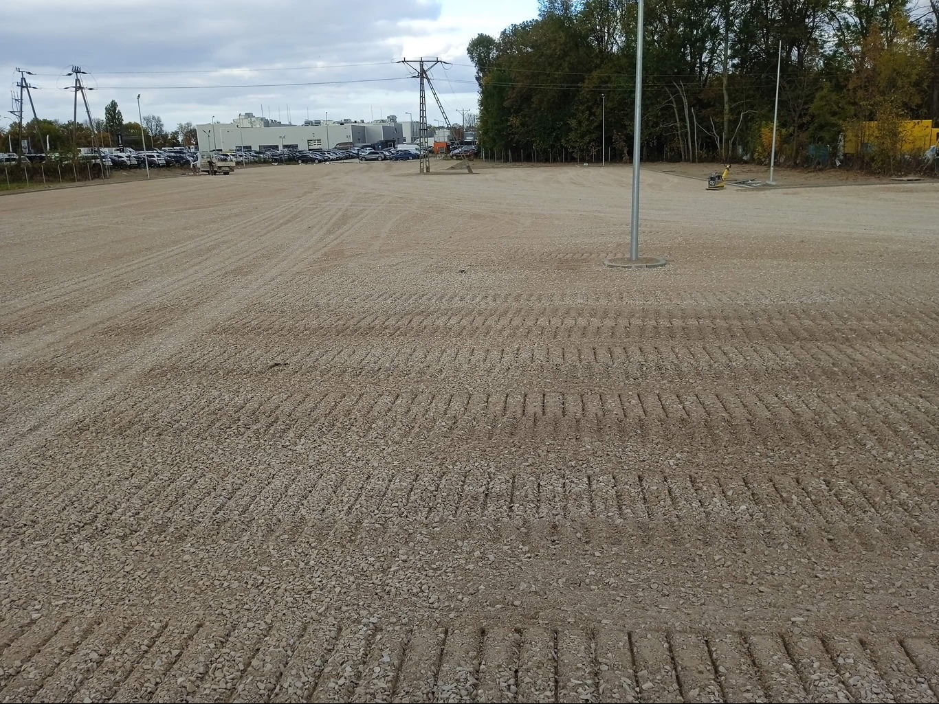 Przygotowany, wyrównany teren pod budowę parkingu w Kobyłce, z widocznymi śladami po walcu i słupami oświetleniowymi w tle.