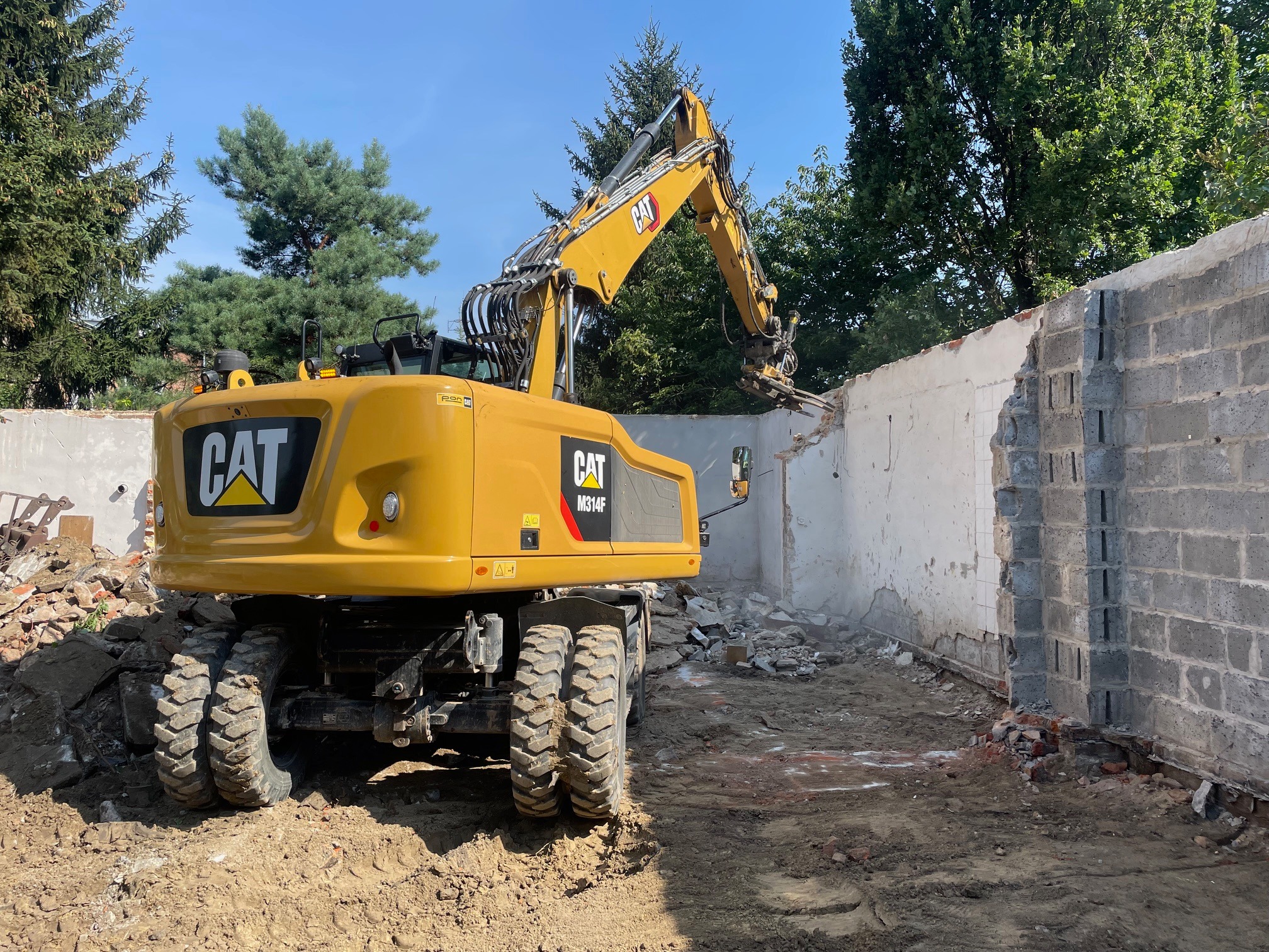 Żółta koparka kołowa marki CAT M314F w trakcie rozbiórki murowanej ściany, widok z tyłu, z gruzem na ziemi i drzewami w tle.