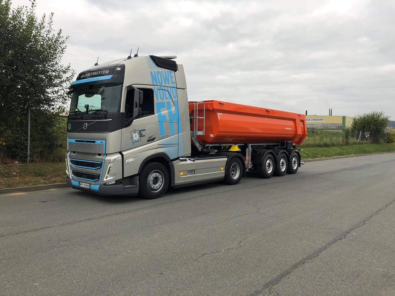 Ciężarówka Volvo FH z pomarańczową naczepą wywrotką, zaparkowana na asfaltowej drodze, z widocznym budynkiem przemysłowym w tle.