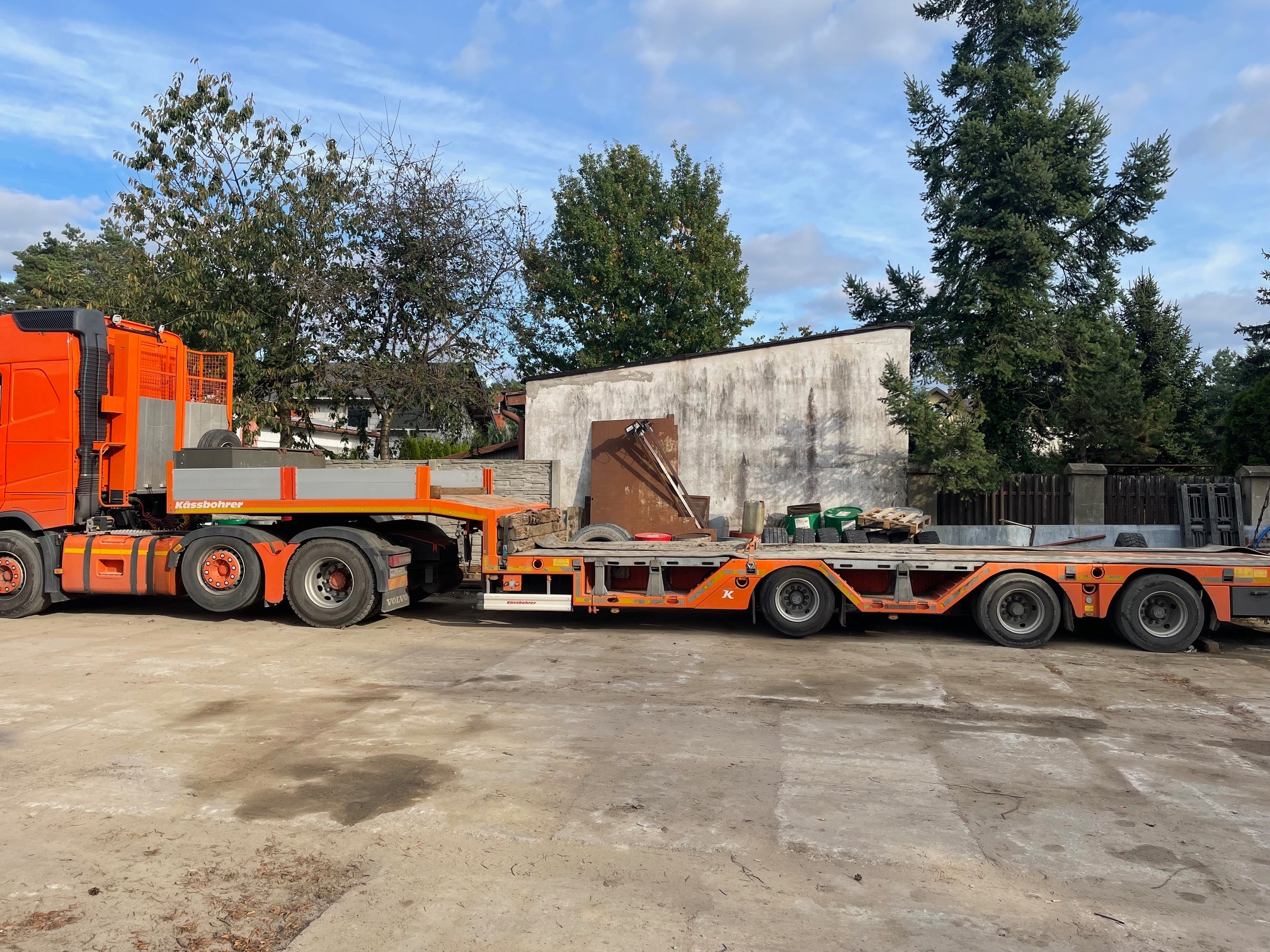 Pomarańczowa ciężarówka z niskopodwoziową naczepą Kässbohrer na betonowym placu, z widocznymi drzewami i budynkiem w tle.