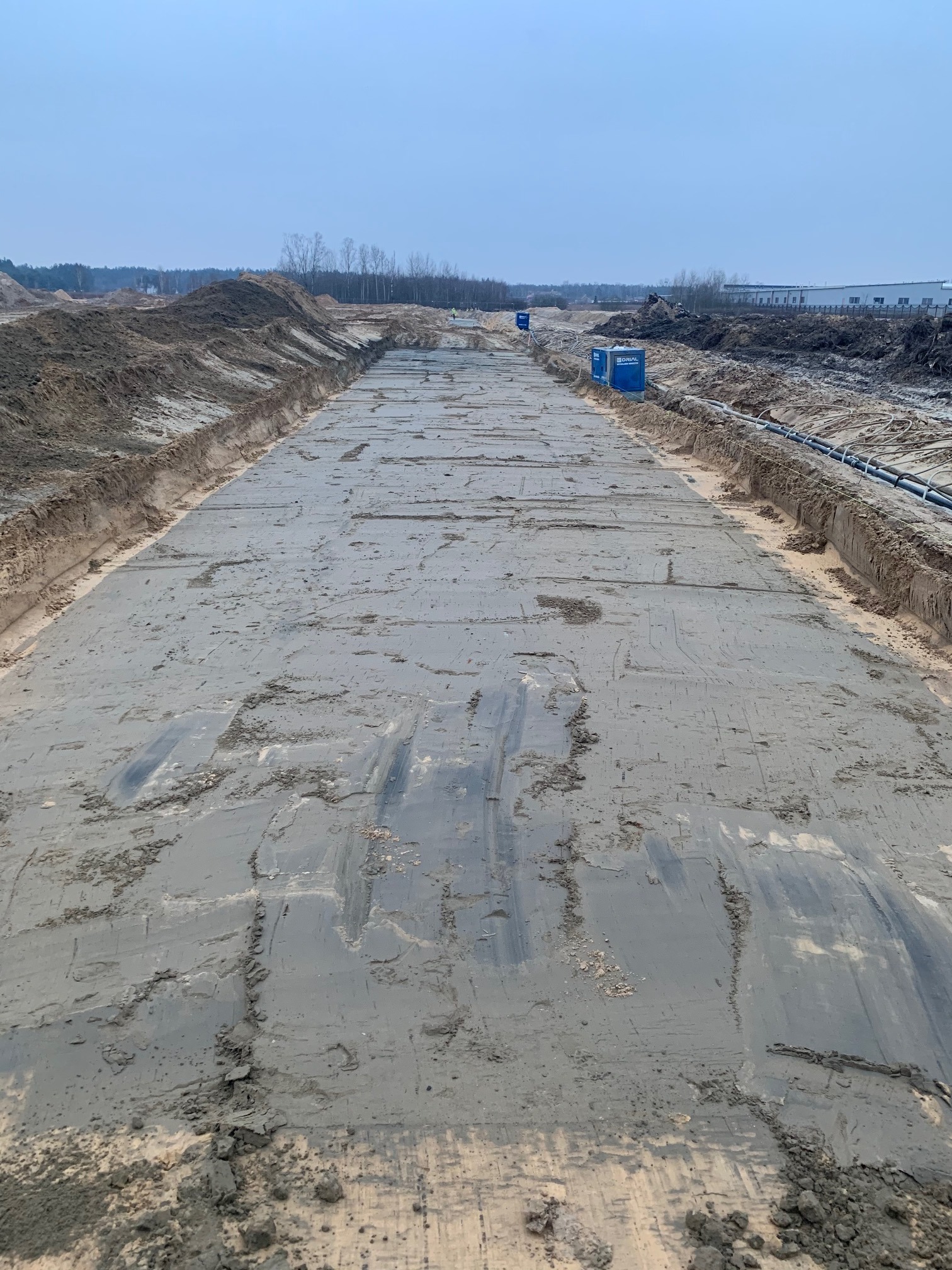 Przygotowany teren pod budowę drogi, z widocznymi śladami maszyn i wyrównaną powierzchnią, w tle zabudowania i linia drzew.