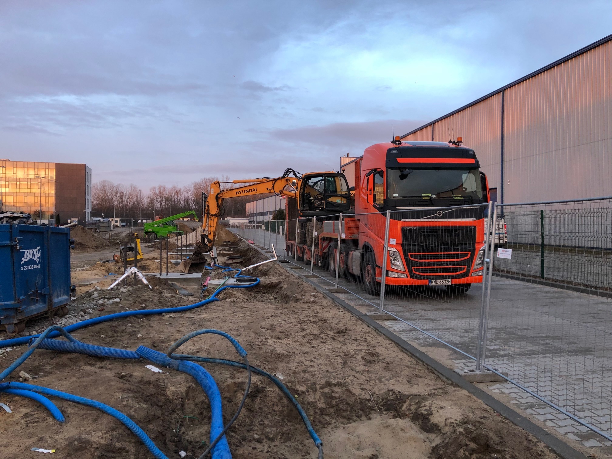 Pomarańczowy ciągnik siodłowy Volvo przewozi żółtą koparkę Hyundai na niskopodwoziowej naczepie, widoczne niebieskie rury na ziemi i teren budowy w tle.