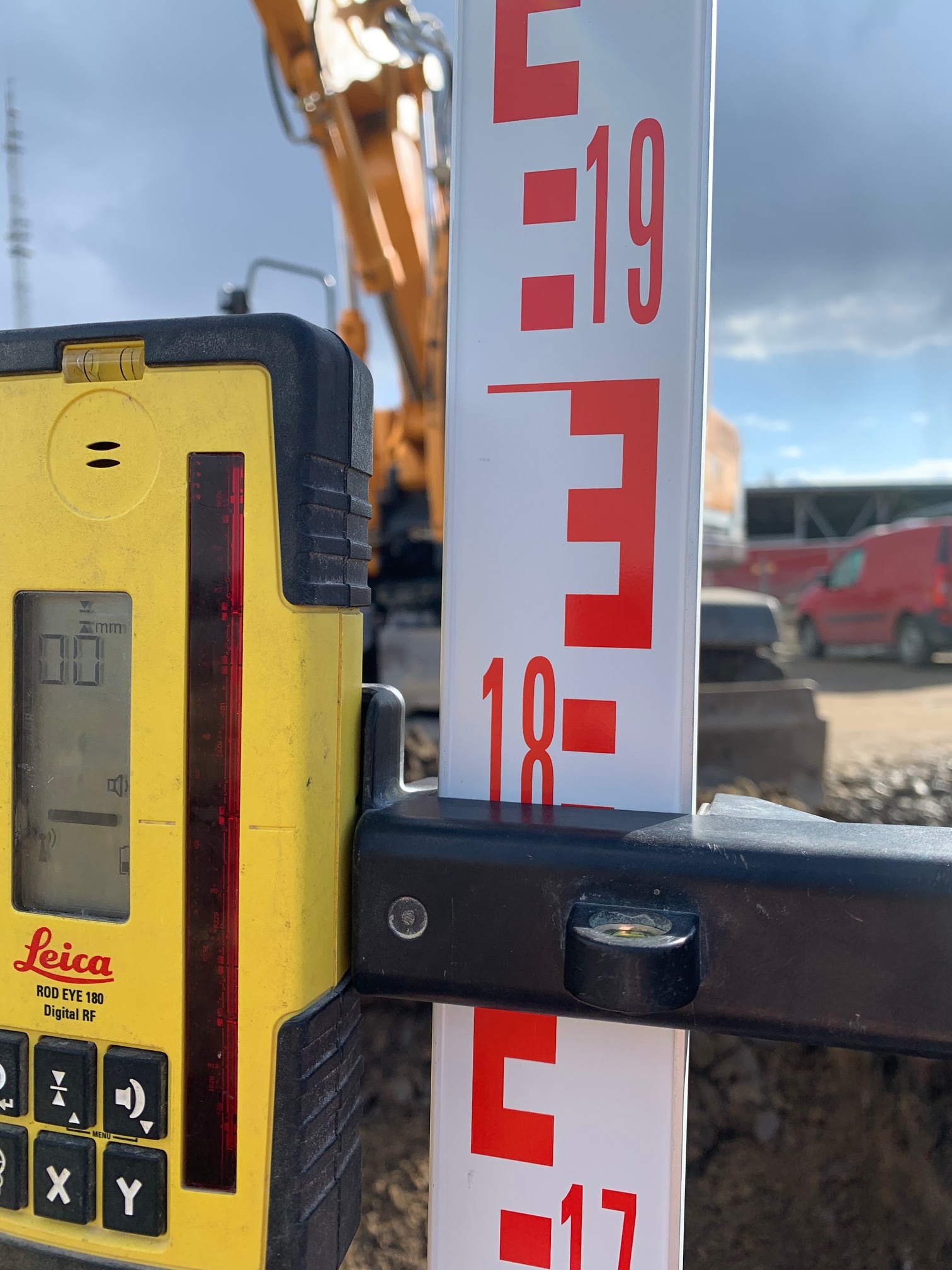 Żółty niwelator Leica Rod Eye 180 przymocowany do łaty niwelacyjnej z czerwonymi oznaczeniami, w tle koparka i samochód dostawczy na placu budowy.