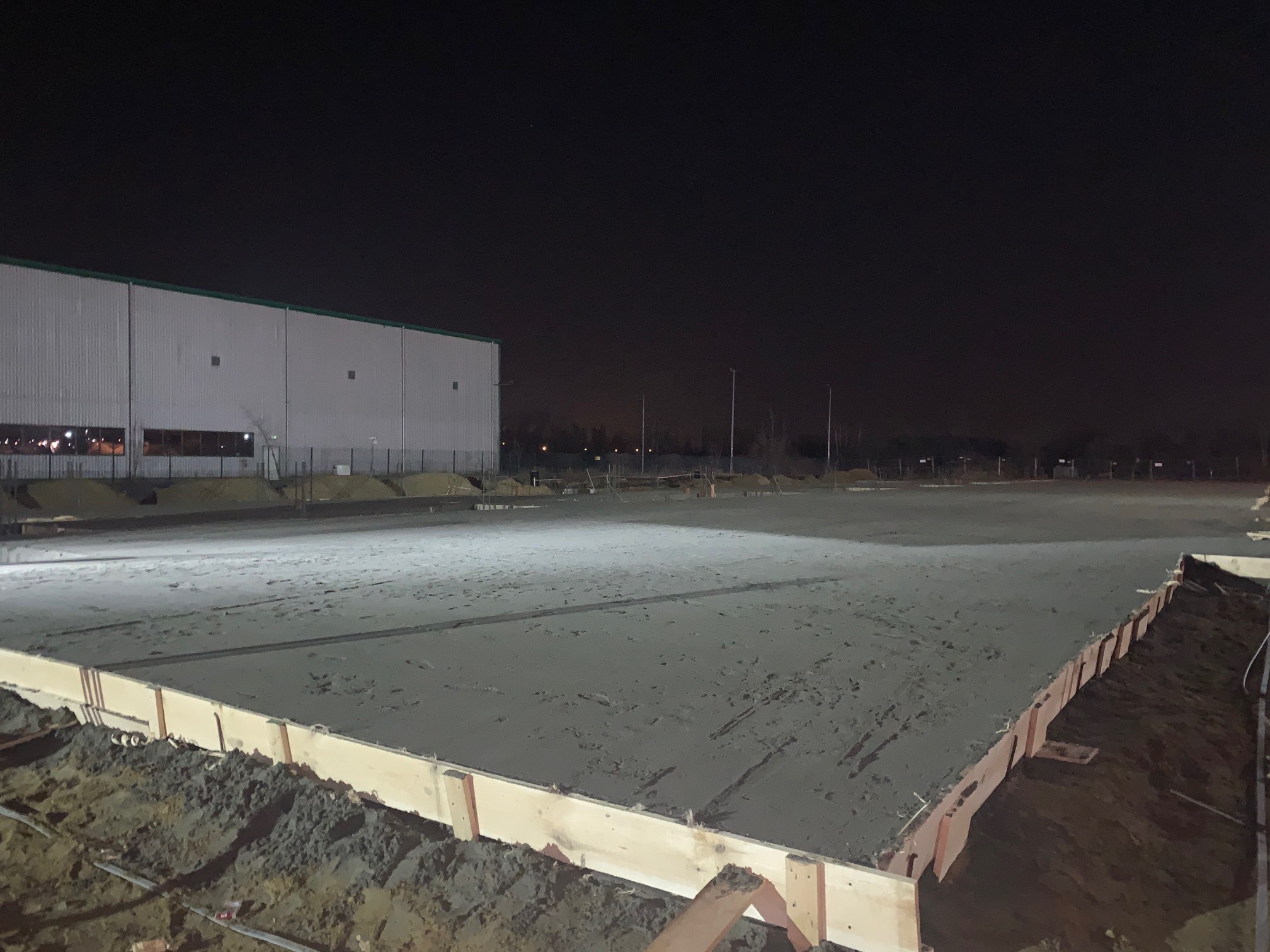 Wylana płyta betonowa w drewnianym szalunku na placu budowy nocą, w tle budynek magazynowy.