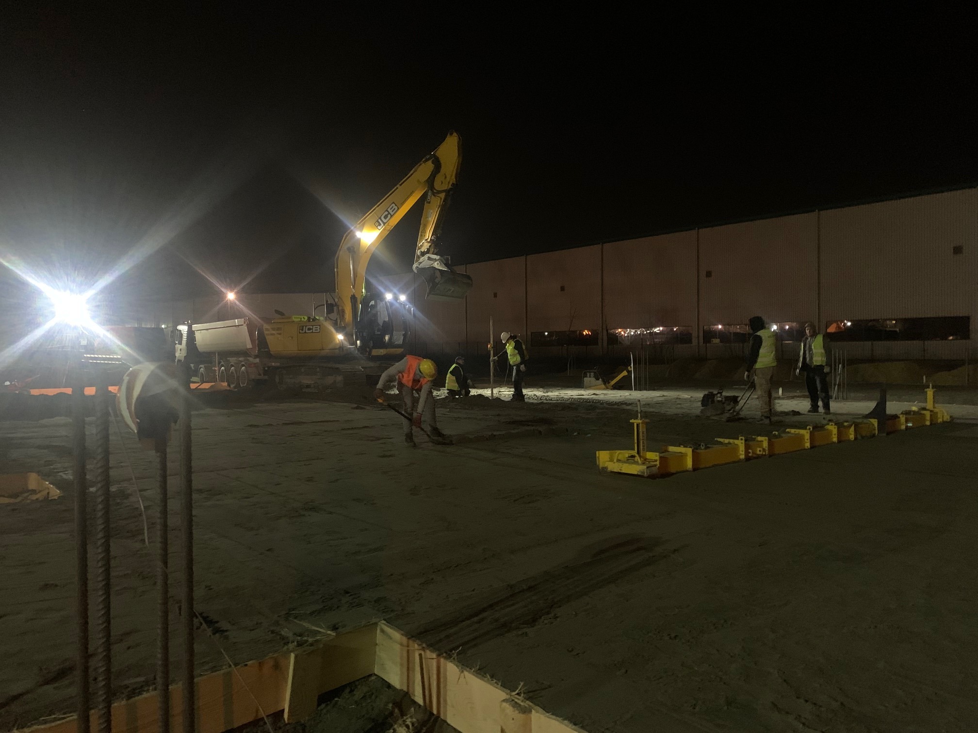 Nocna niwelacja terenu pod budowę, widoczna koparka JCB, pracownicy w kamizelkach odblaskowych i przygotowane zbrojenie.