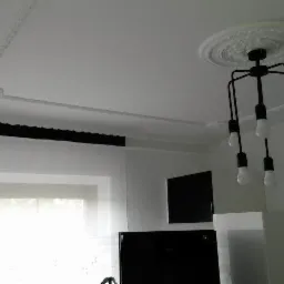 Biały sufit z dekoracyjną sztukaterią i nowoczesną czarną lampą wiszącą z żar&oacute;wkami, widok na okno i ścianę z telewizorem.