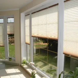 Jasne, plisowane osłony okienne zamontowane w białych oknach z widokiem na zielony ogród. Wnętrze z roślinami doniczkowymi.