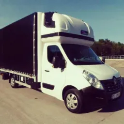 Biały Renault Master z czarną plandeką na pace, zaparkowany na betonowym placu w słoneczny dzień.