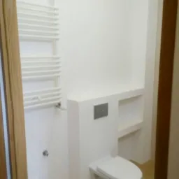 Nowoczesna łazienka z podwieszaną toaletą w zabudowie, grzejnikiem drabinkowym i półką w ścianie, wykończona w bieli i beżu.