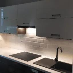 Nowoczesna kuchnia z białymi szafkami, czarnym zlewem i płytą indukcyjną, podświetlane płytki ścienne typu metro, lodówka ozdobiona magnesami.