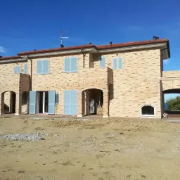 Dwupiętrowy dom z cegły z niebieskimi okiennicami i otwartymi drzwiami balkonowymi, w trakcie budowy, z widokiem na morze w oddali, Campofilone, Włochy.