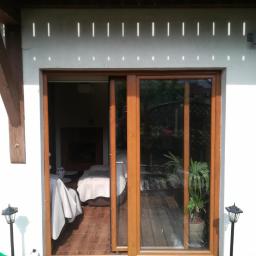 Finestre Okna Sp. z o.o. - Drewniane okno przesuwne z widokiem na wnętrze domu i taras, oświetlone promieniami słońca padającymi przez drewnianą pergolę.