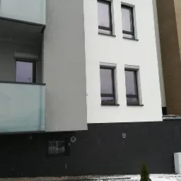 Nowoczesny budynek wielorodzinny z oknami z czarnymi ramami i roletami zewnętrznymi, częściowo otynkowany na biało i szaro, z balkonem z mlecznego szkła, widziany w zimowy dzień podczas opadów...