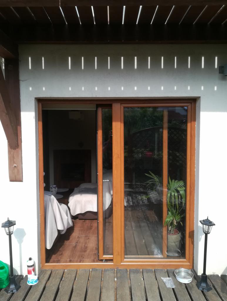 Drewniane okno przesuwne z widokiem na wnętrze domu i taras, oświetlone promieniami słońca padającymi przez drewnianą pergolę.