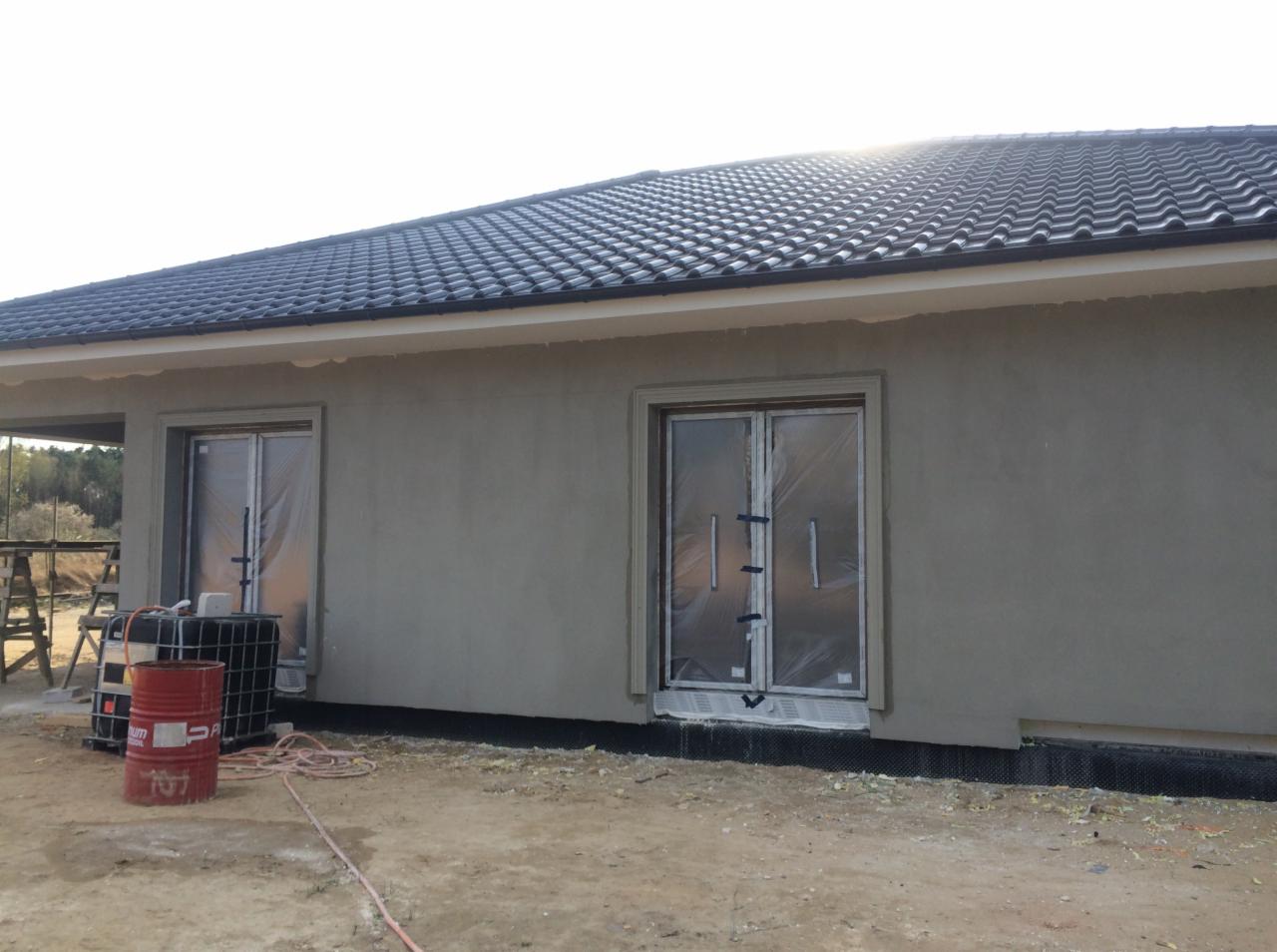 Budynek w trakcie prac wykończeniowych: elewacja otynkowana na szaro, okna zabezpieczone folią, czarny dach z dachówki ceramicznej, widoczne materiały budowlane i przewód na ziemi.
