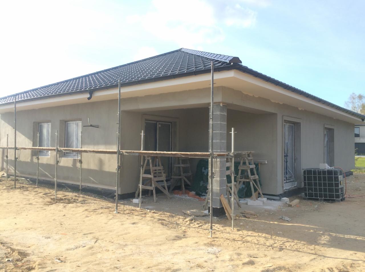 Dom jednorodzinny w trakcie tynkowania zewnętrznego, widoczne rusztowania i niezabezpieczone okna.