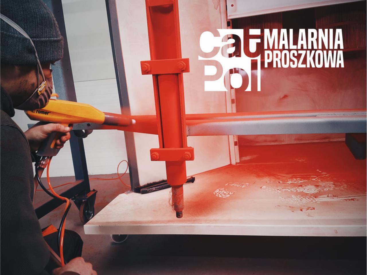 Pracownik w masce i czapce maluje proszkowo metalowy element na czerwono w kabinie lakierniczej, używając pistoletu Romer.