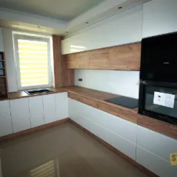 Nowoczesna kuchnia z białymi szafkami i drewnianymi blatami, z zabudowanym piekarnikiem i płytą indukcyjną, oraz oknem z żółtymi żaluzjami.