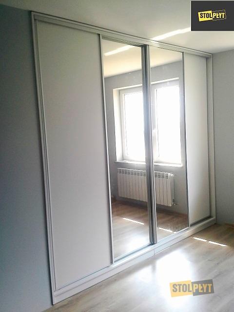 Biała szafa przesuwna z lustrami odbijającymi okno i kaloryfer, widoczny fragment podłogi z paneli.