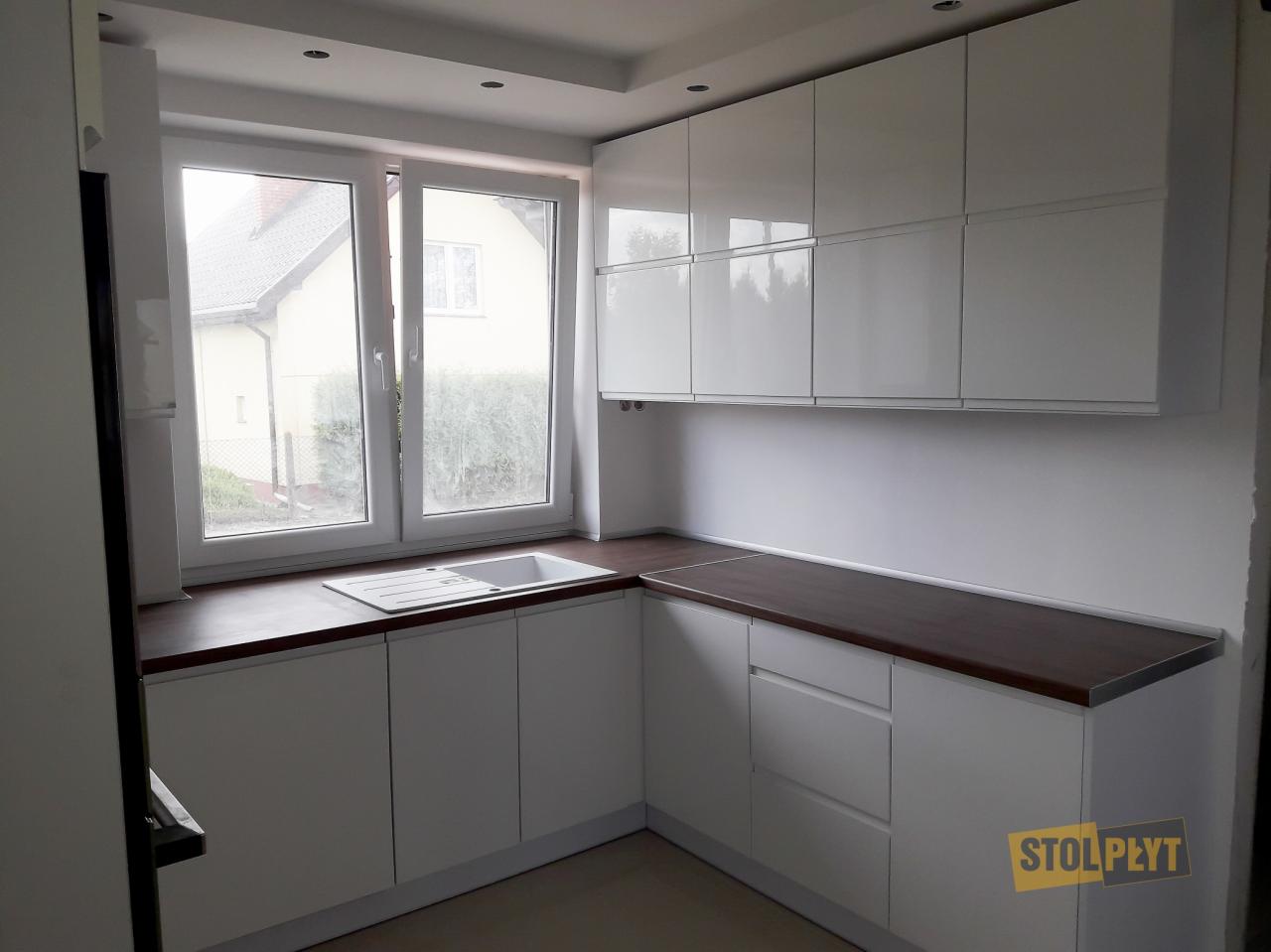 Nowoczesna, biała zabudowa kuchenna z ciemnym blatem, widok na okno z zewnątrz, logo firmy Stolpłyt w prawym dolnym rogu.