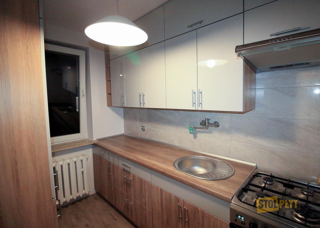 Jasna kuchnia z drewnianym blatem i białymi szafkami, widok na okno i grzejnik, oświetlenie sufitowe w stylu nowoczesnym.