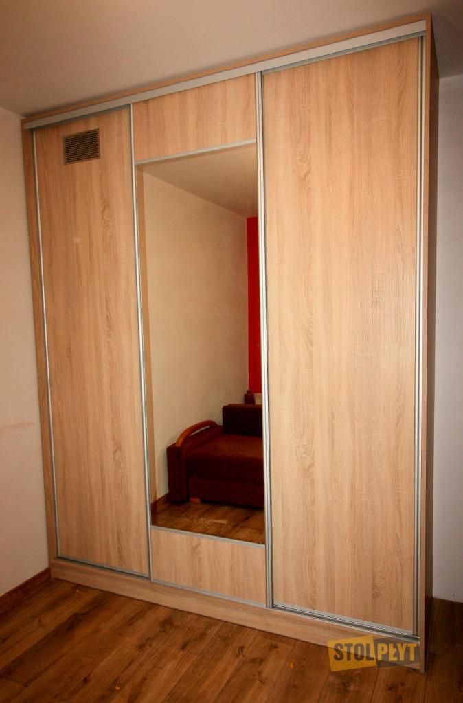 Trzydrzwiowa szafa przesuwna z lustrem, okleina drewnopodobna w ciepłym odcieniu, widoczny fragment sofy odbity w lustrze.