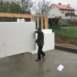 Montaż paneli izolacyjnych na drewnianej konstrukcji domu w trakcie budowy, widoczne białe panele z charakterystycznym wzorem łączeń, fragmenty zbrojonej betonowej wylewki i dom w tle.