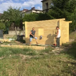 Dwóch mężczyzn buduje drewnianą konstrukcję w ogrodzie, jeden z łopatą, drugi z miotłą, widoczne deski i narzędzia.