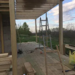 Wnętrze w trakcie budowy tarasu z widokiem na zewnątrz, widoczne dwie drabiny, narzędzia i materiały budowlane.