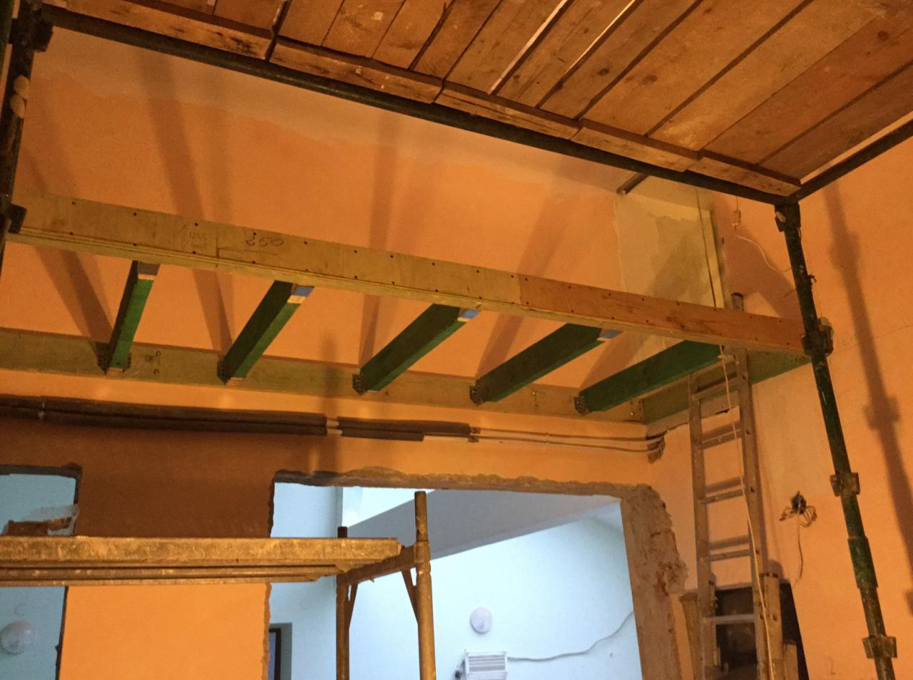 Wnętrze w trakcie remontu, widoczne drewniane belki podtrzymujące strop, metalowe rusztowanie i drabina oparta o ścianę, ściany w odcieniach beżu i pomarańczy, widoczny otwór drzwiowy do innego...