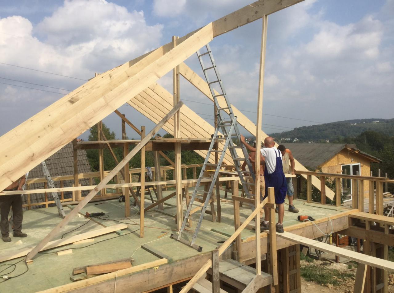 Konstrukcja drewniana domu w trakcie budowy, widoczni pracownicy na drabinie, w tle zielone wzgórza i częściowo wykończony budynek z żółtą elewacją.