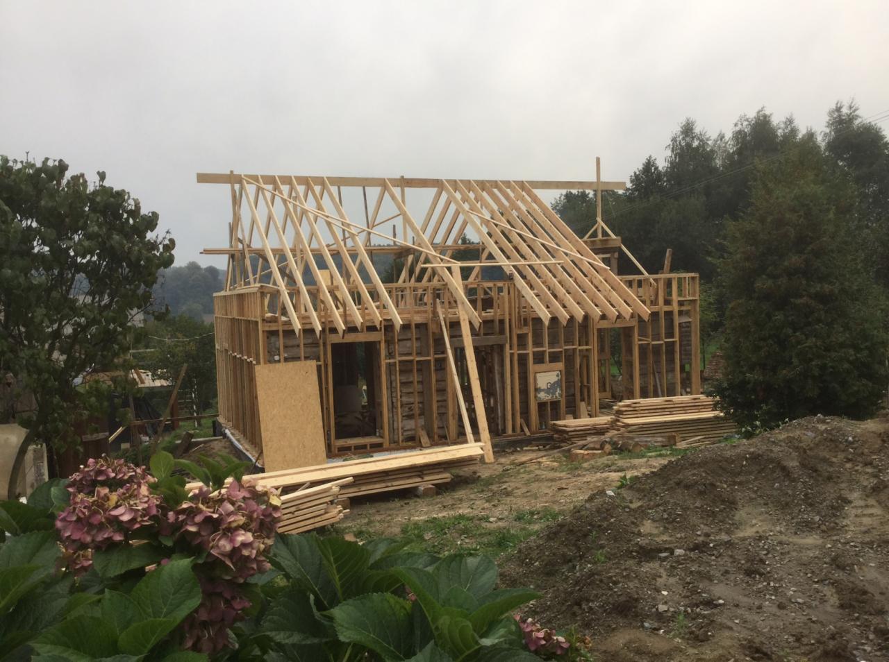 Konstrukcja domu w trakcie budowy: drewniany szkielet z widocznymi elementami więźby dachowej, stos desek obok, kwiaty hortensji na pierwszym planie, pochmurne niebo w tle.