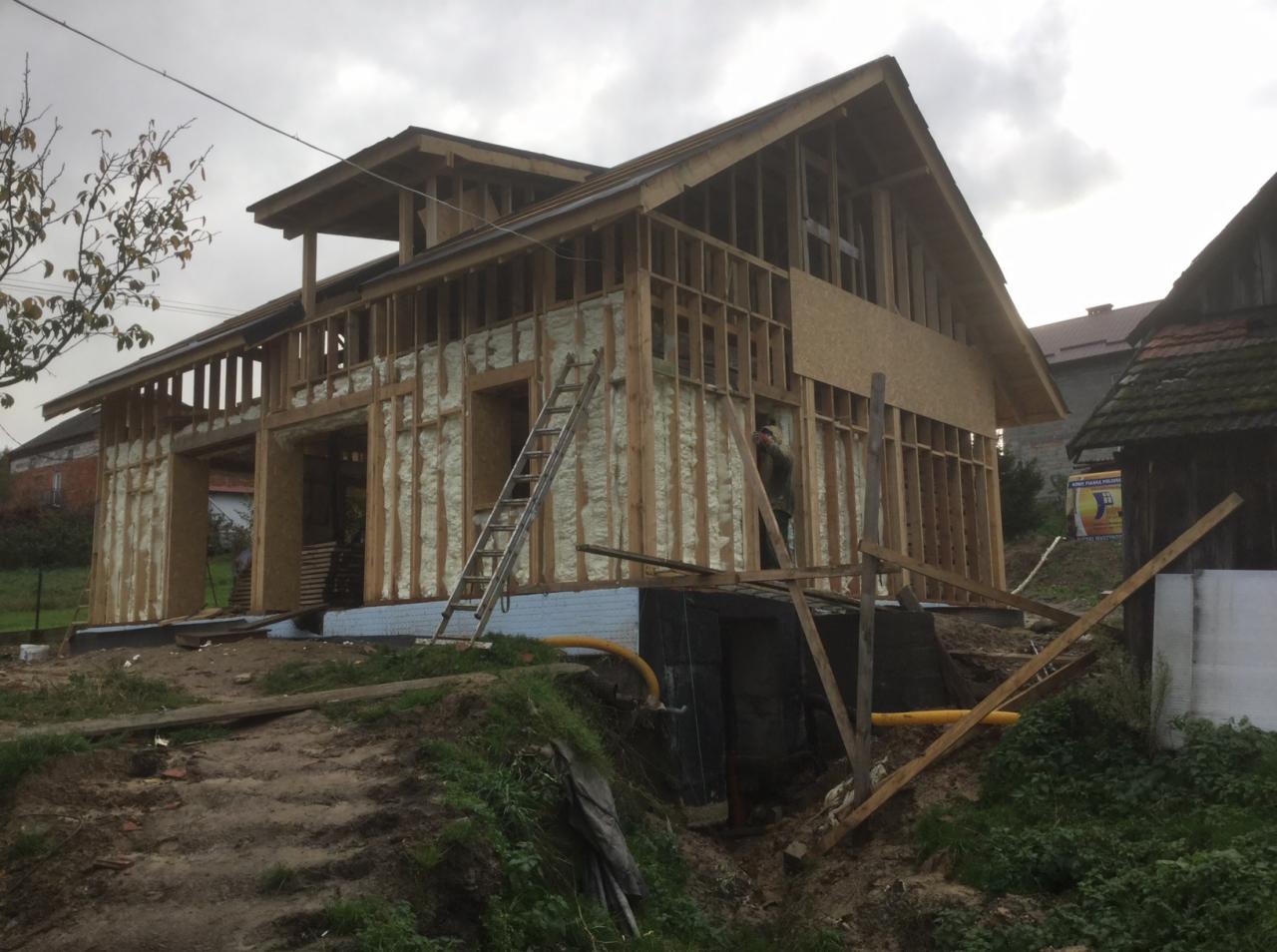 Budowa domu szkieletowego w trakcie: widoczna drewniana konstrukcja, ocieplenie pianą, drabina oparta o ścianę, pracownik wewnątrz konstrukcji.