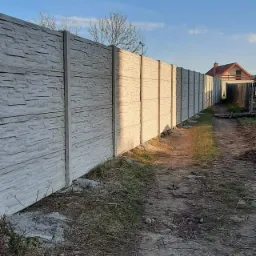 Długi, szary, betonowy płot z fakturą kamienia, z widocznym domem w tle i ścieżką wzdłuż ogrodzenia.