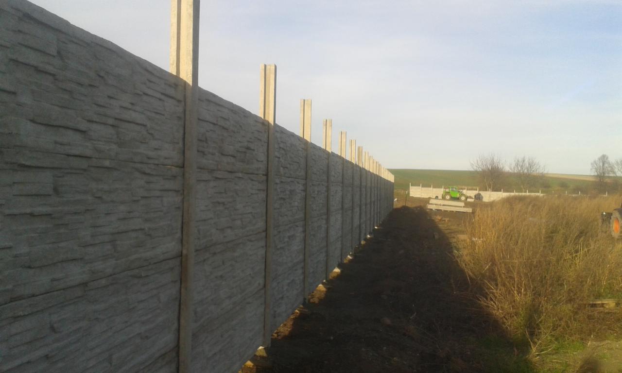 Długi, szary mur z elementów imitujących kamień, z betonowymi słupkami, ciągnący się wzdłuż pola, z widocznym traktorem w tle, w słoneczny dzień.